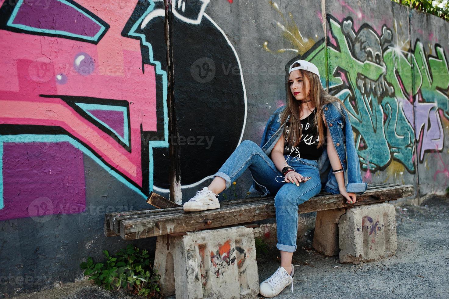 elegante chica hipster casual con gorra y jeans usa música para escuchar desde auriculares de teléfono móvil contra una gran pared de graffiti con bomba. foto