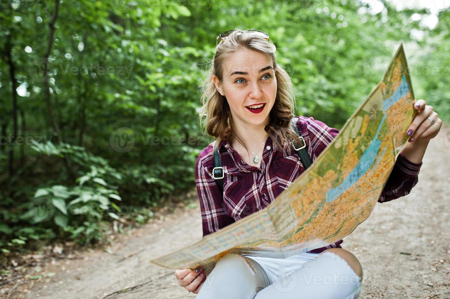 retrato de una joven y hermosa rubia positiva sentada en el suelo con un mapa en las manos en el bosque. foto