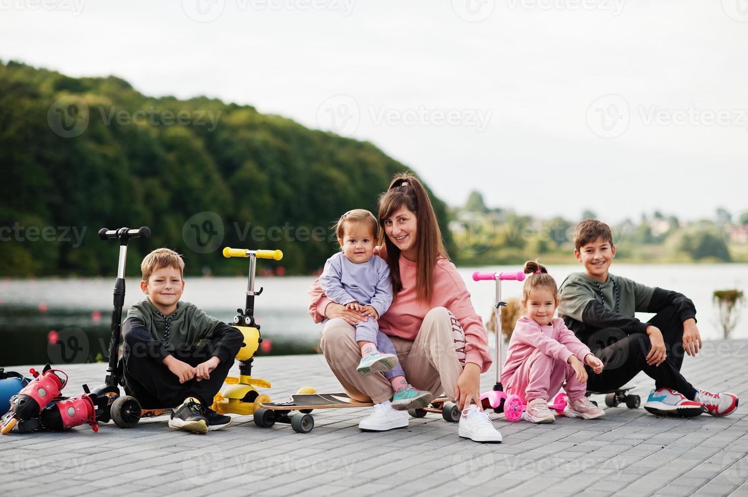 joven madre elegante con cuatro hijos al aire libre. la familia deportiva pasa tiempo libre al aire libre con scooters y patines. foto