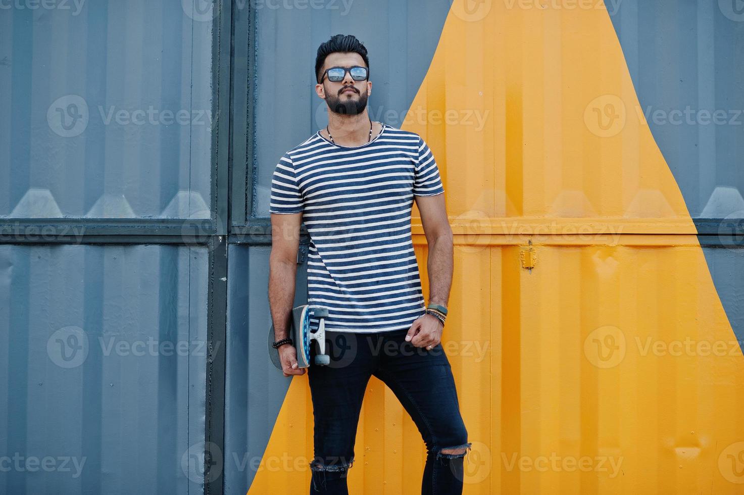 Handsome tall arabian beard man model at stripped shirt posed outdoor. Fashionable arab guy at sunglasses with skateboard against yellow painted wall. photo