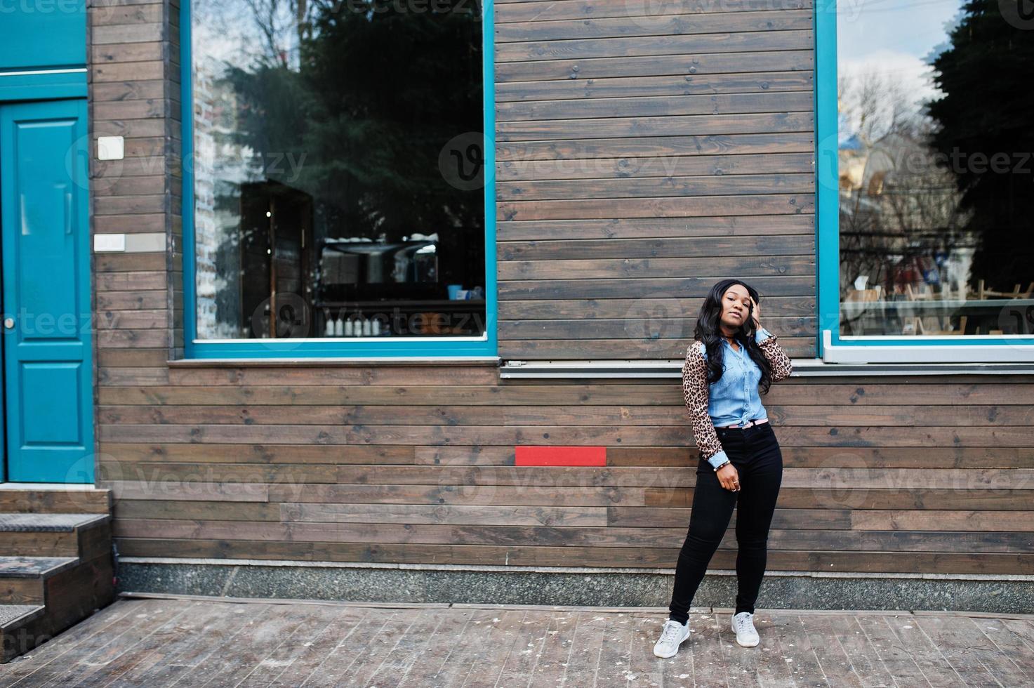 chica afroamericana hipster con camisa de jeans con mangas de leopardo posando en la calle contra una casa de madera con ventanas. foto