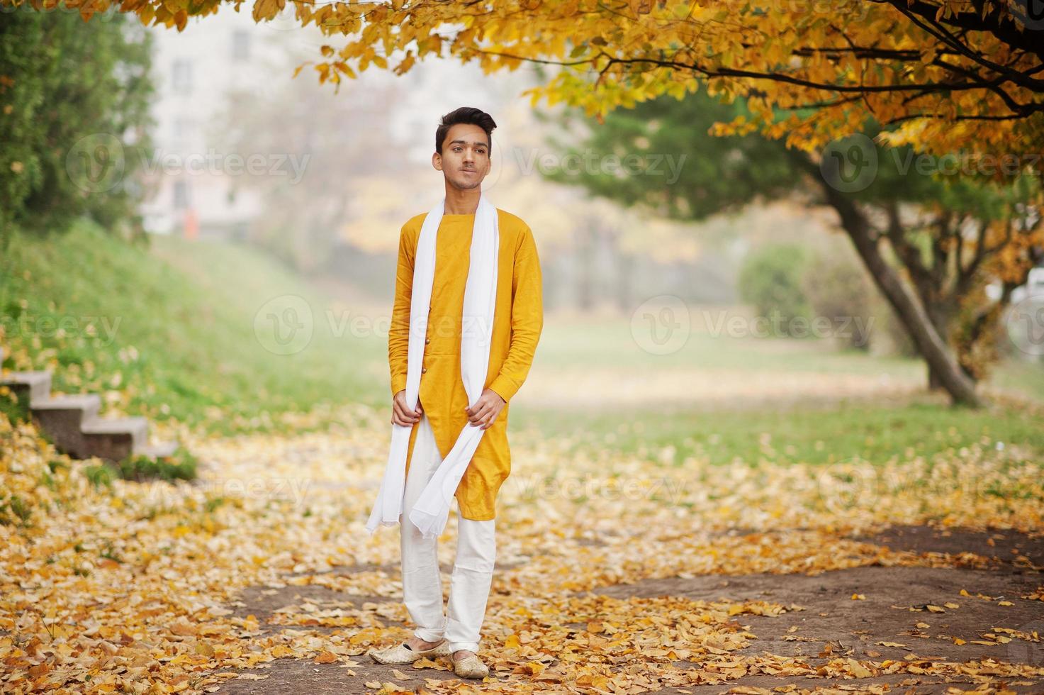 hombre indio con estilo en ropa tradicional amarilla con bufanda blanca posada al aire libre contra el árbol de hojas de otoño. foto