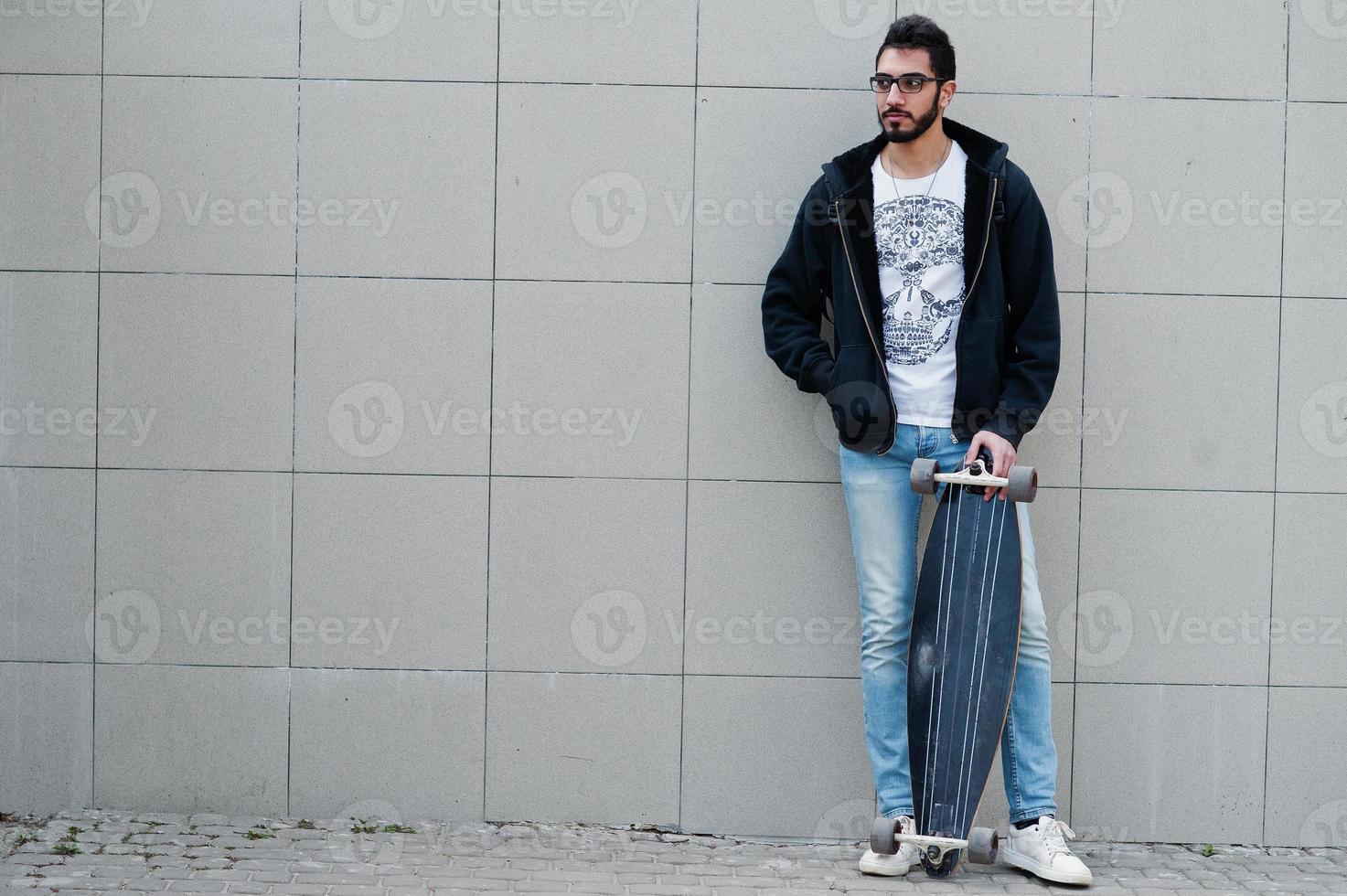 Street style arab man in eyeglasses with longboard posed against gray wall. photo