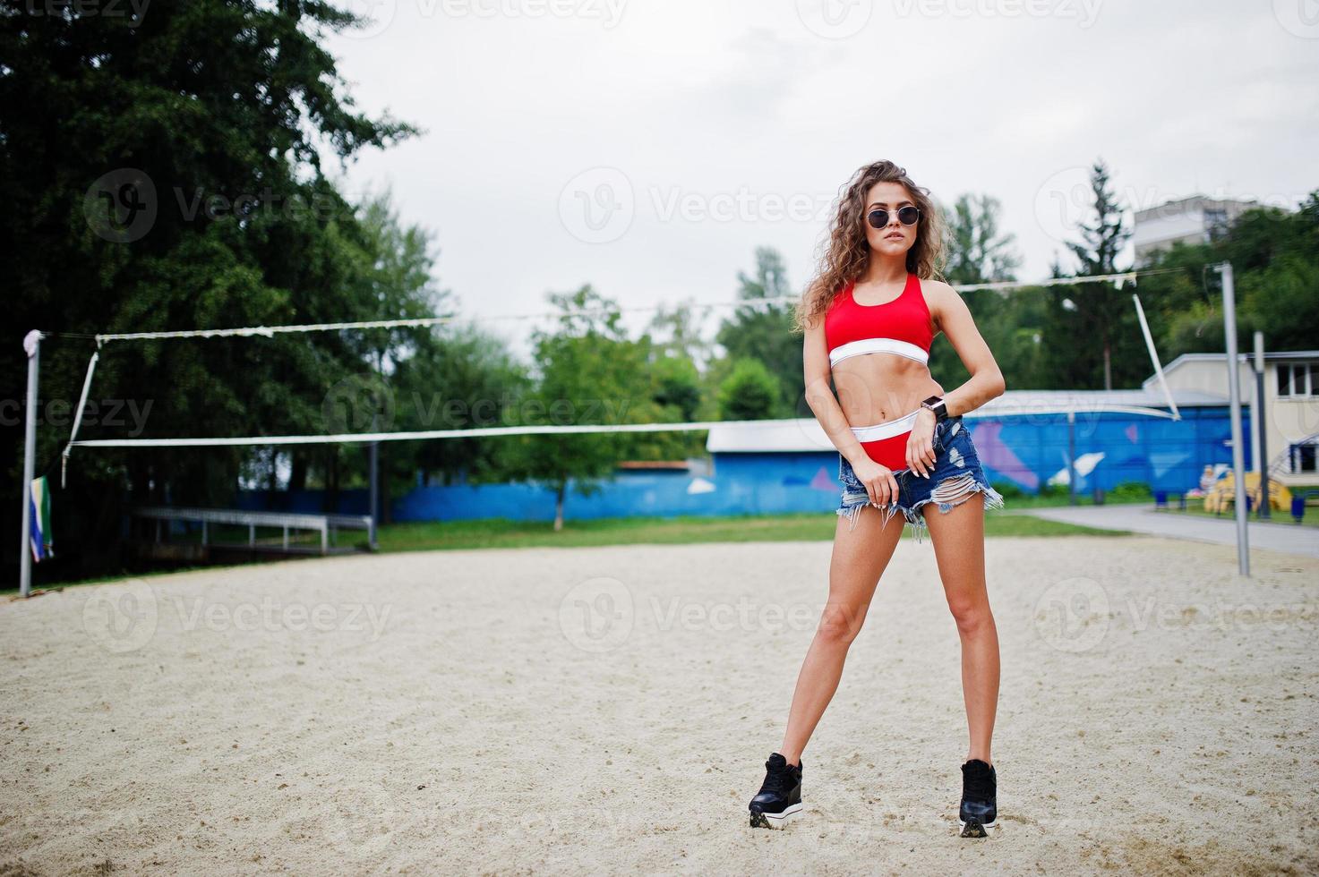 chica sexy modelo rizada en top rojo, pantalones cortos de mezclilla, copa y gafas de sol posadas en el campo de voleibol de playa. foto
