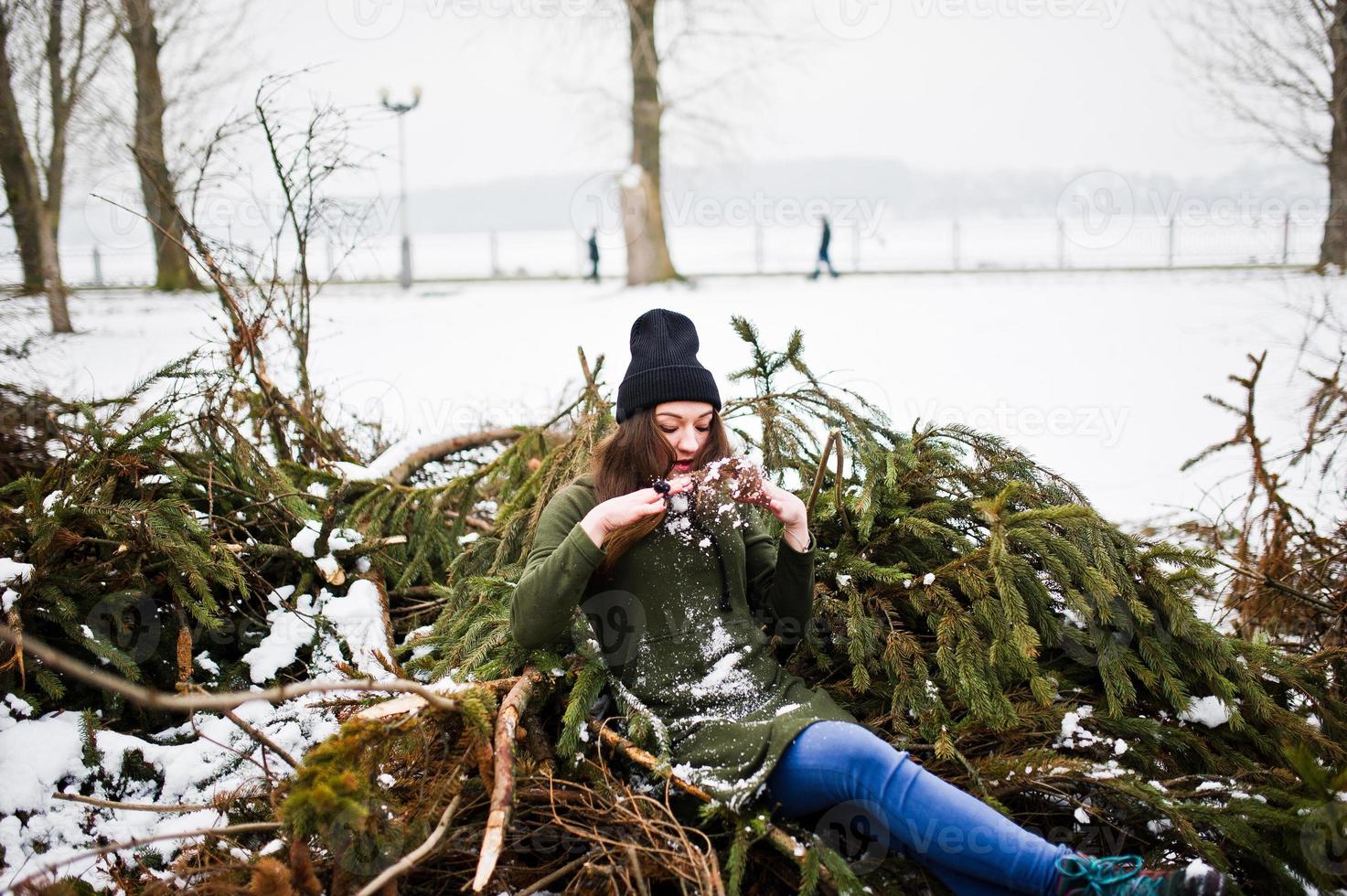 Young girl wear on long green sweatshirt, jeans and black headwear at  branches of the pine tree in winter day. photo