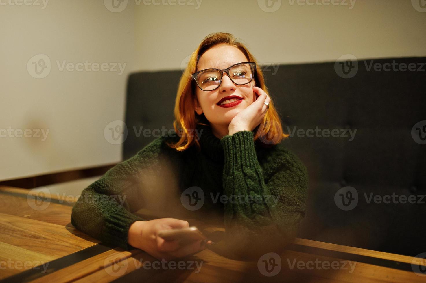 Cheerful young beautiful redhaired woman in glasses, wear on green warm wool sweater,  sitting at cafe and using her mobile phone. photo