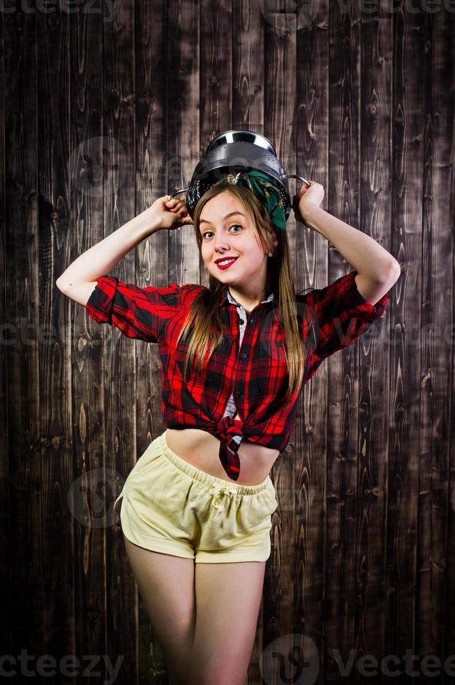 joven ama de casa divertida en camisa a cuadros y pantalones cortos amarillos estilo pin up con tamiz de cocina sobre fondo de madera. foto