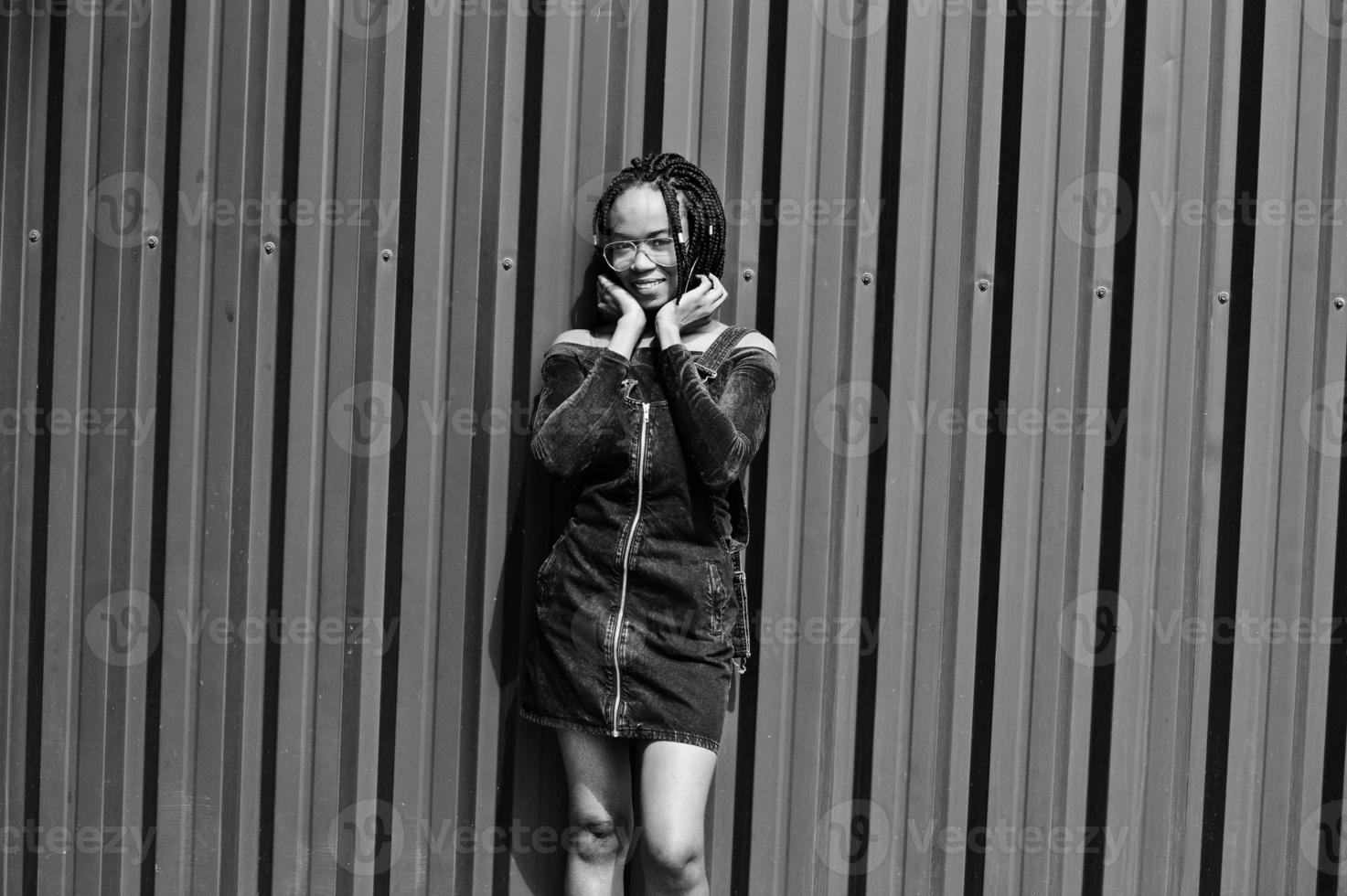 African woman in denim overalls skirt, eyeglasses posed against green steel wall. photo