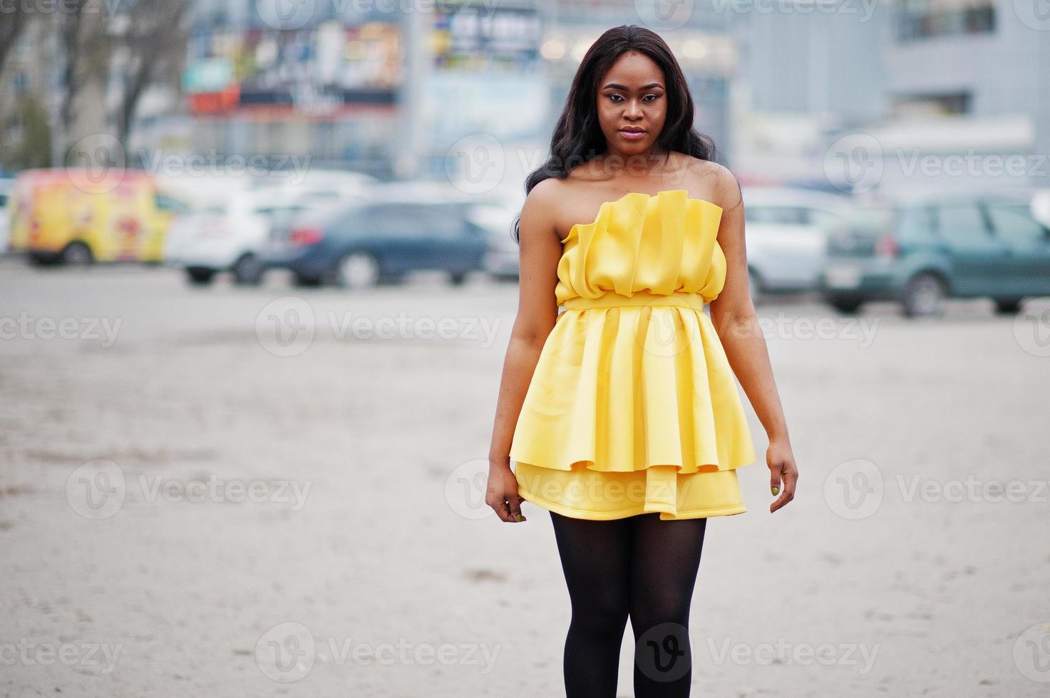 Stylish african american woman at yellow dress posed outdoor car parking. photo