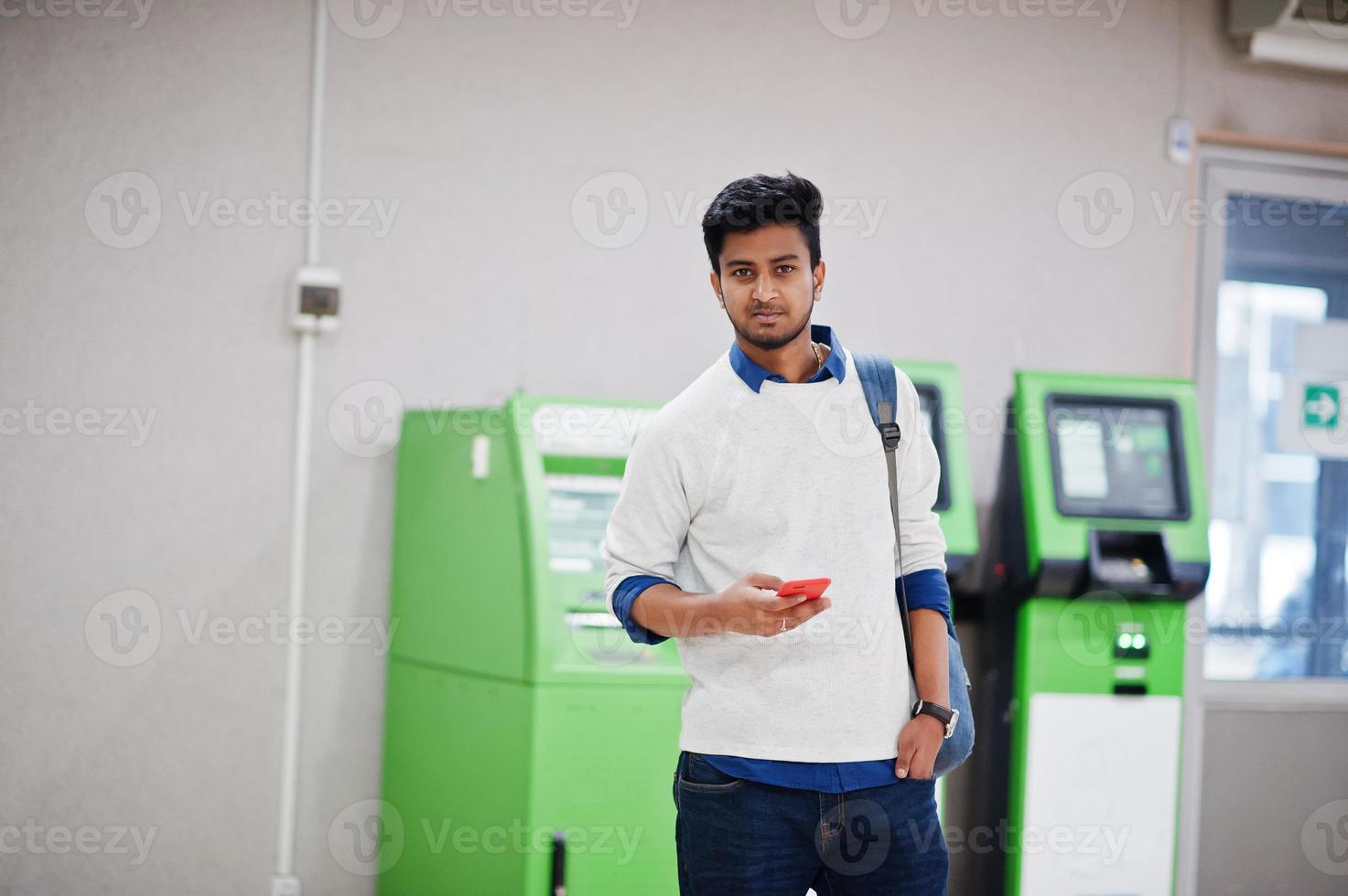 joven asiático con estilo con teléfono móvil y mochila contra la fila de cajero automático verde. foto