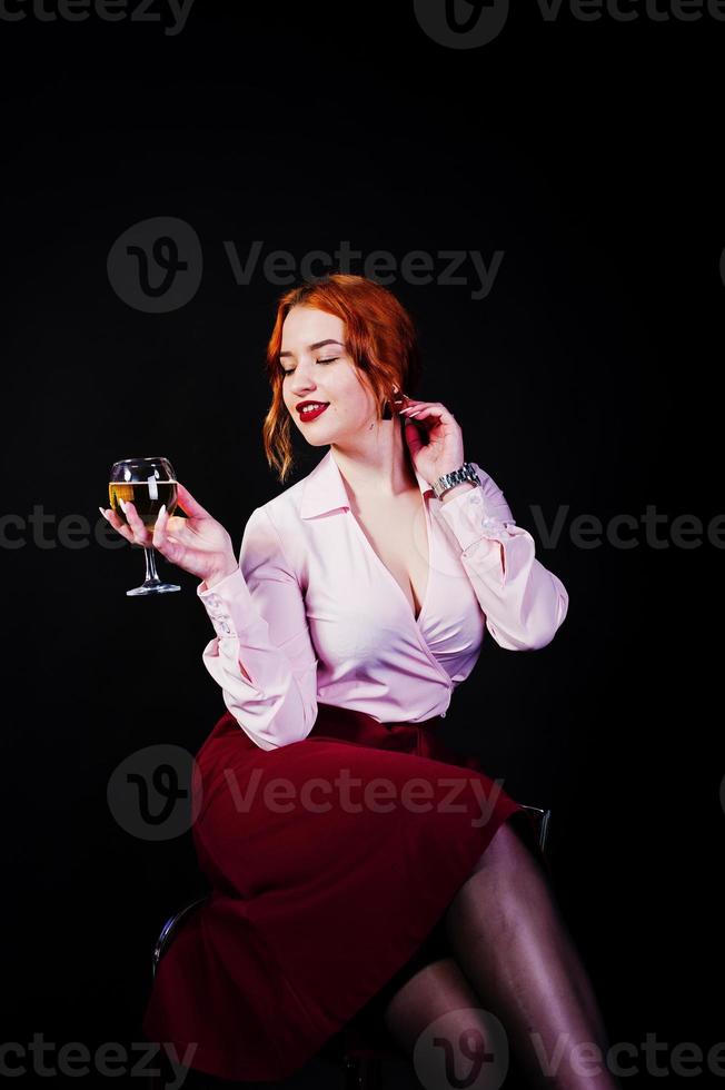 Gorgeous red haired girl in pink blouse and red skirt with glass of wine at hand isolated on black. photo