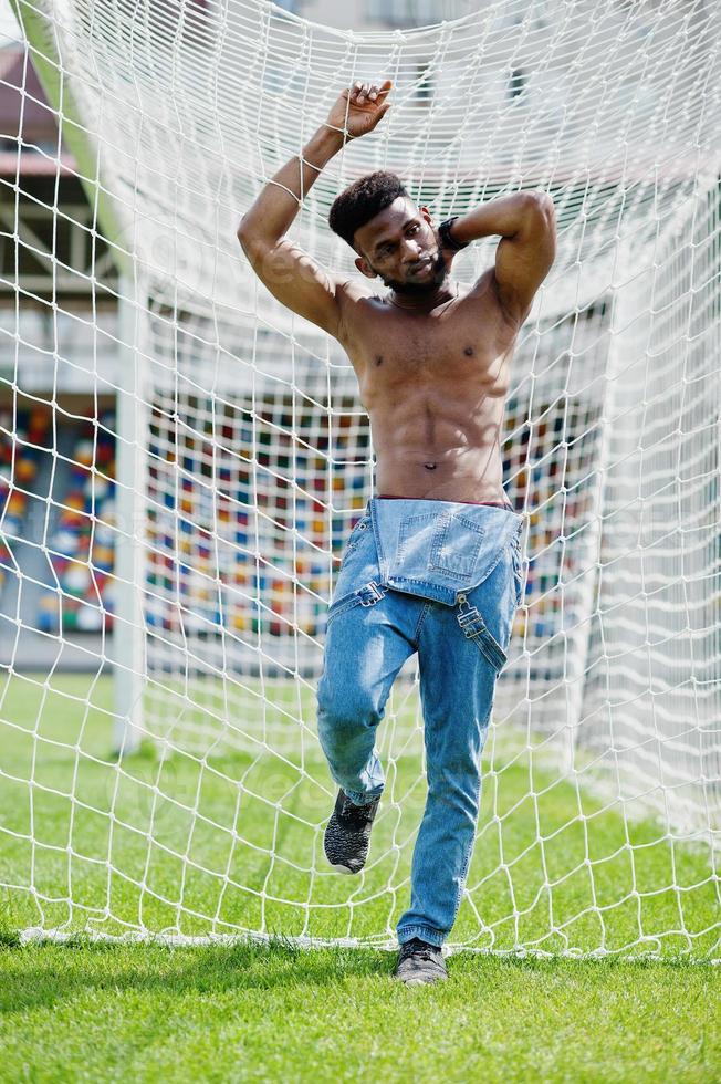 Handsome sexy african american muscular bare torso man at jeans overalls posed at green grass agasinst football gates of stadium field. Fashionable black man portrait. photo