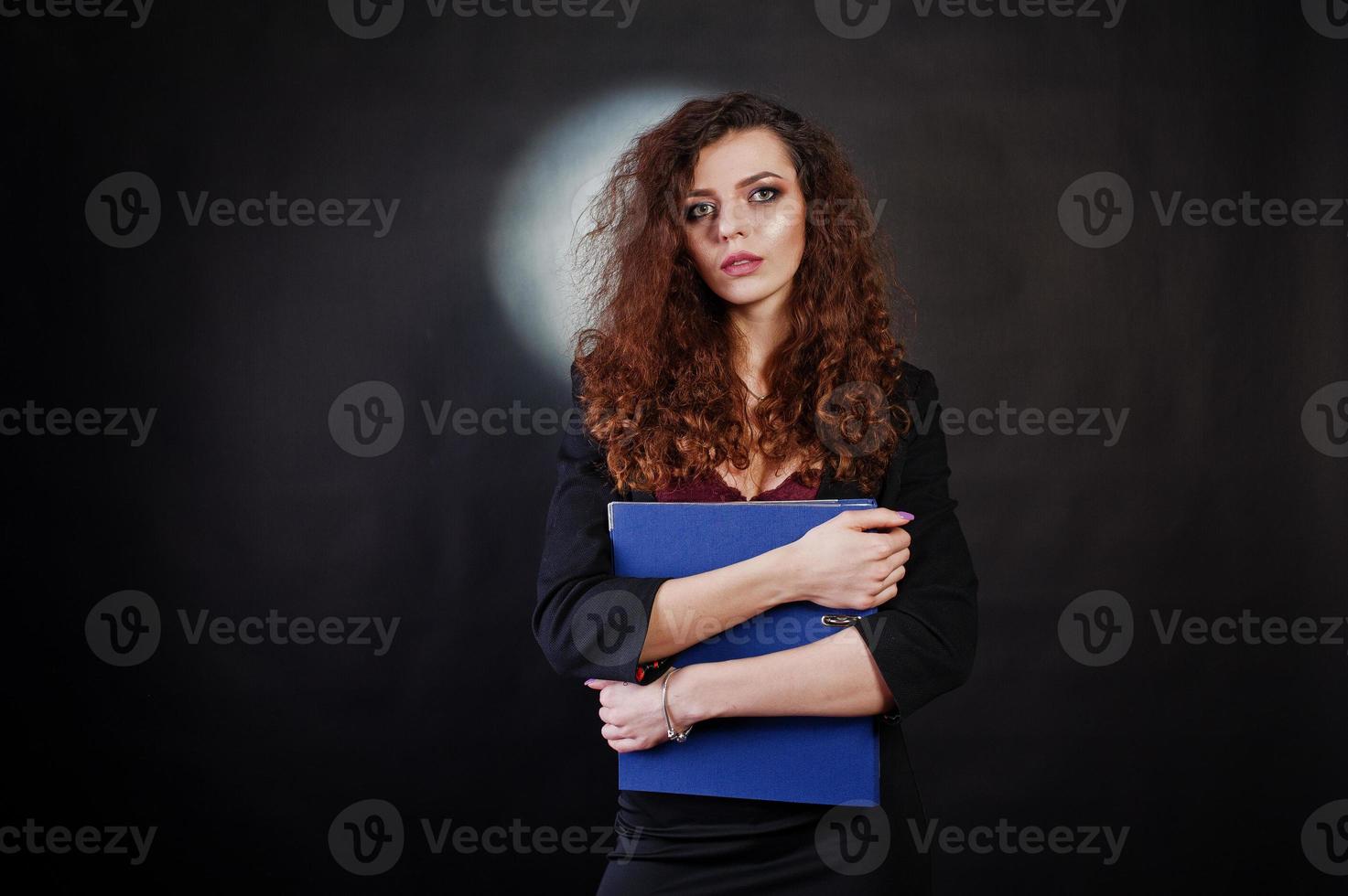 Brunette curly haired girl in black office jacket with skirt, on bra with folder of documentts at hands on studio against black background. Sexy businesswoman. photo