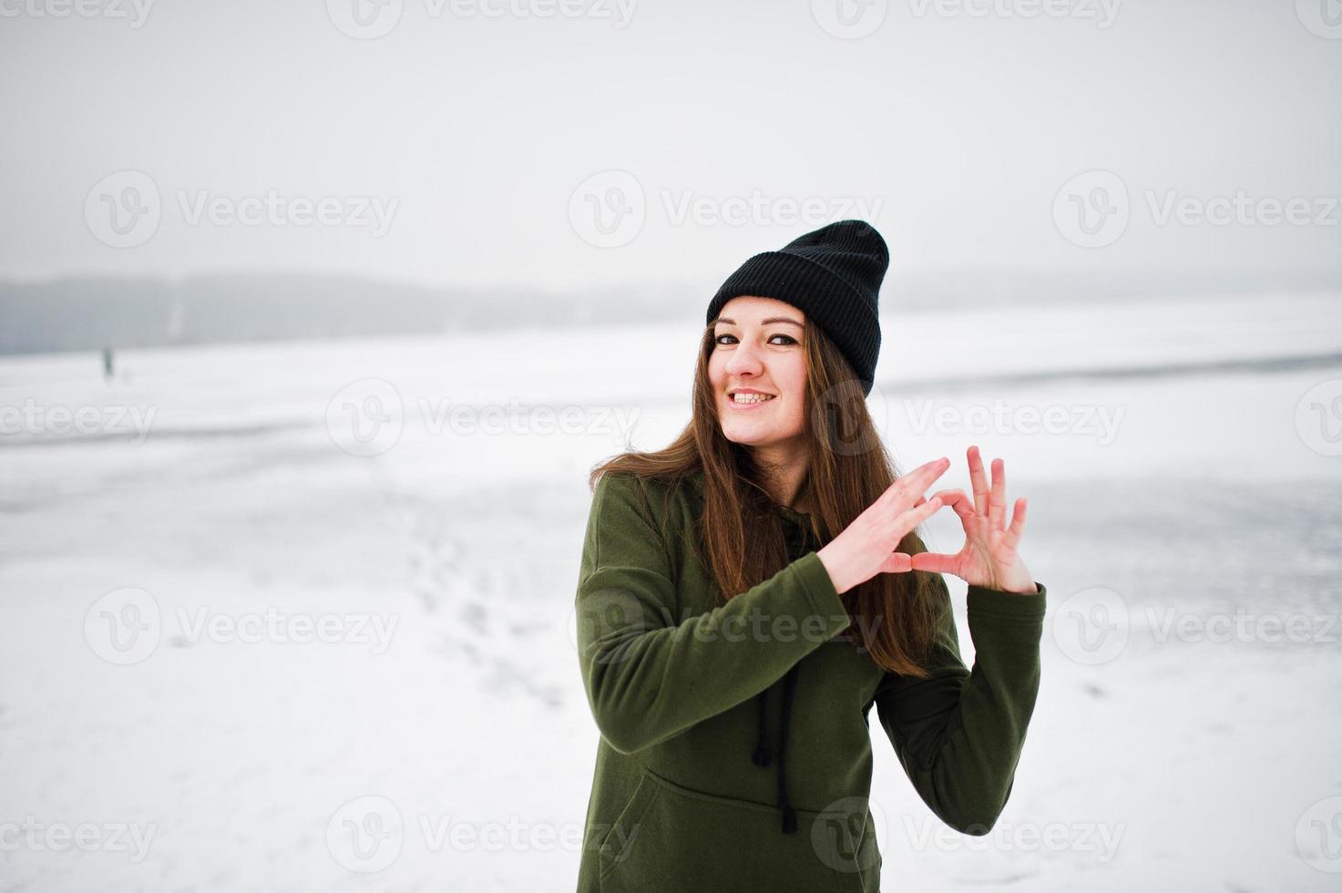 Funny girl wear on long green sweatshirt, jeans and black headwear, at frozen lake in winter day, showing heart on fingers. photo