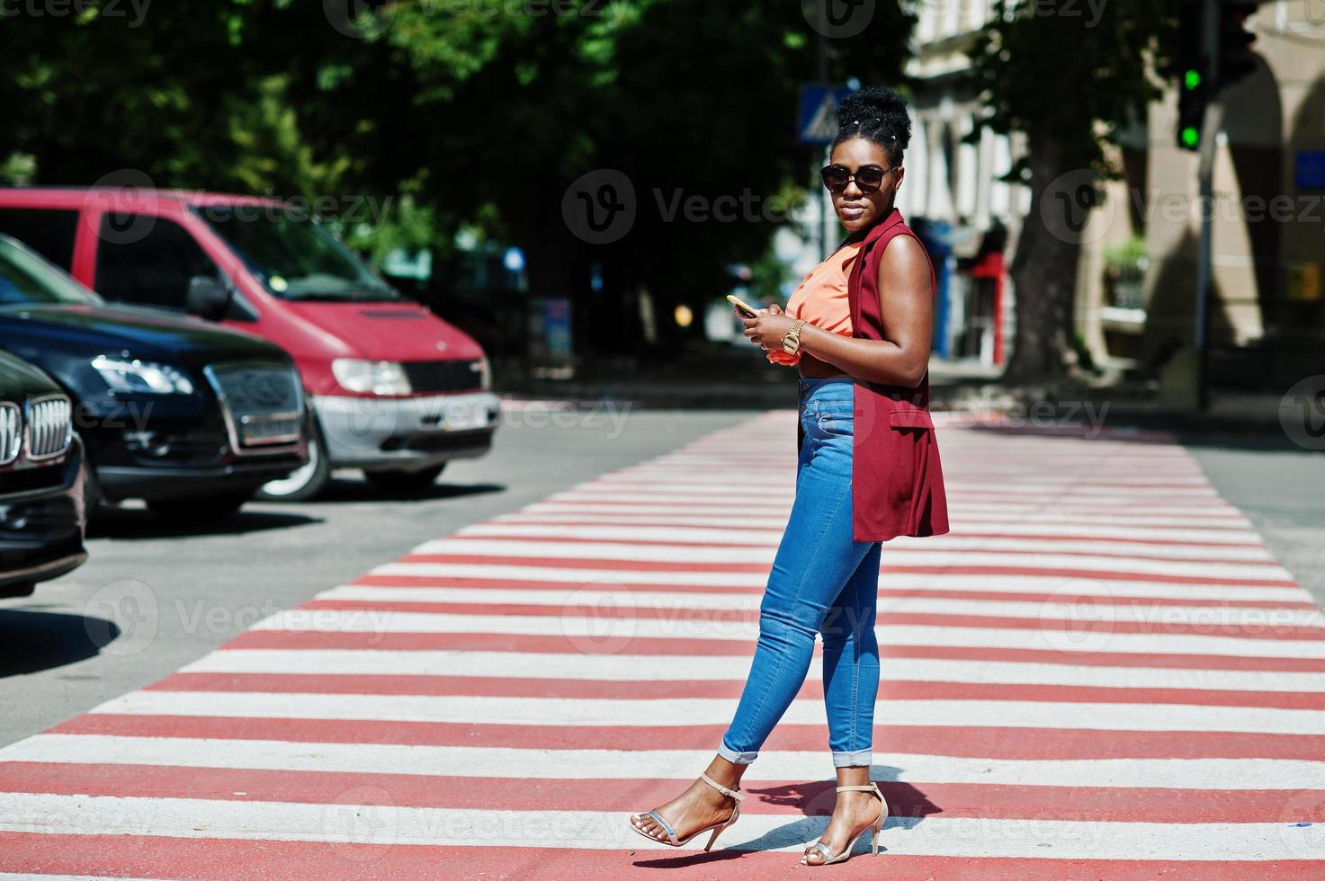 Stylish african american woman walking on crosswalk or pedestrian crossing with mobile phone at hand. photo