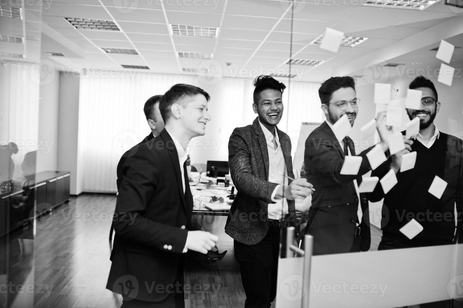 hombre de negocios apuntando sobre vidrio con notas de papel de colores. grupo diverso de empleados varones en ropa formal usando pegatinas. foto