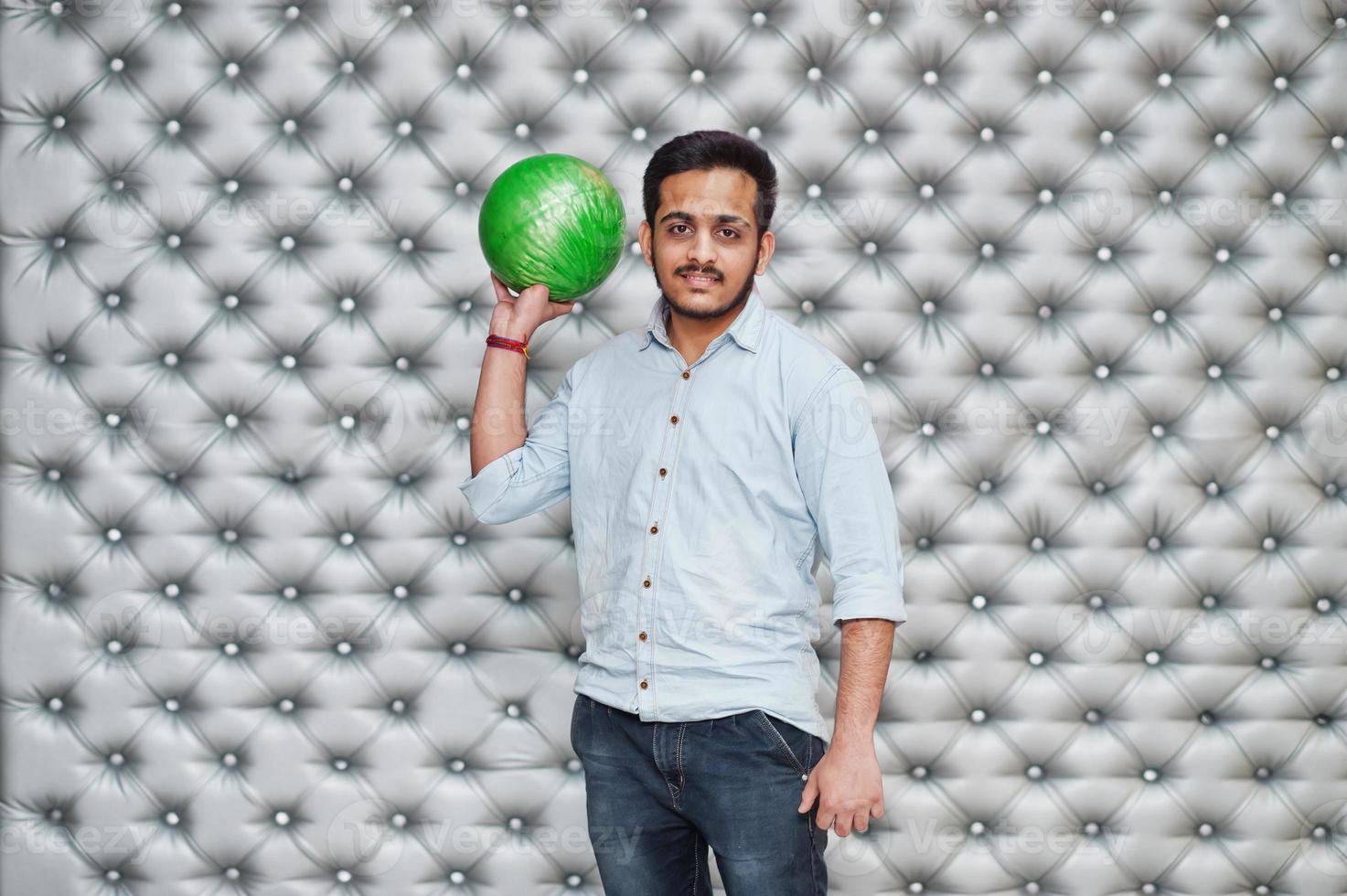 Stylish asian man in jeans shirt standing with bowling ball at hand against silver wall background. photo