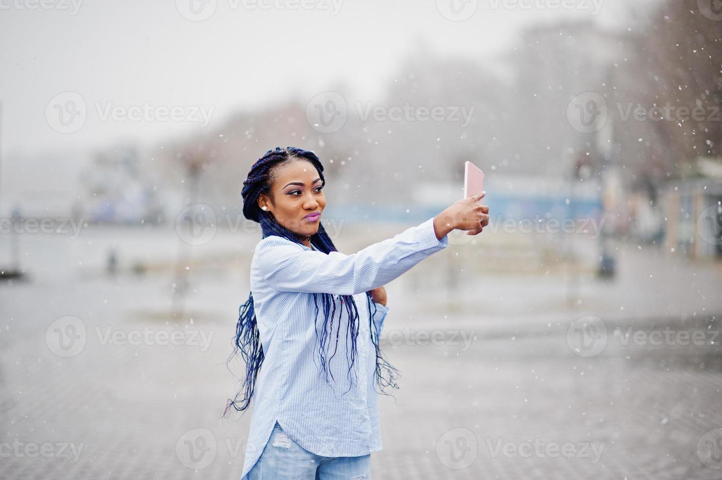 Stylish african american girl with dreads holding mobile phone at hand and making selfie, outdoor with snowy weather. photo