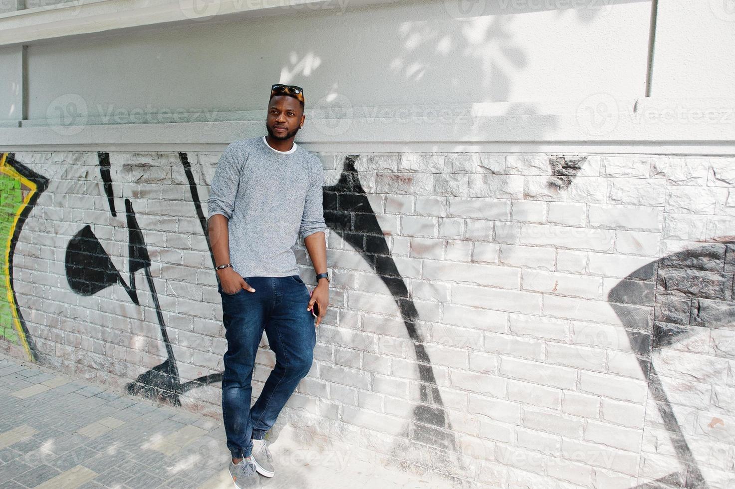 Stylish african american boy on gray sweater and black sunglasses posed at street. Fashionable black guy against graffity wall. photo