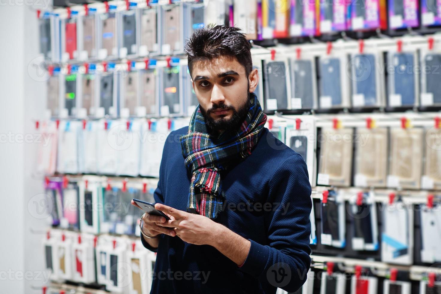 Comprador de cliente de hombre de barba india en la tienda de teléfonos móviles mirando en su teléfono inteligente. concepto de pueblos y tecnologías del sur de Asia. tienda de celulares foto
