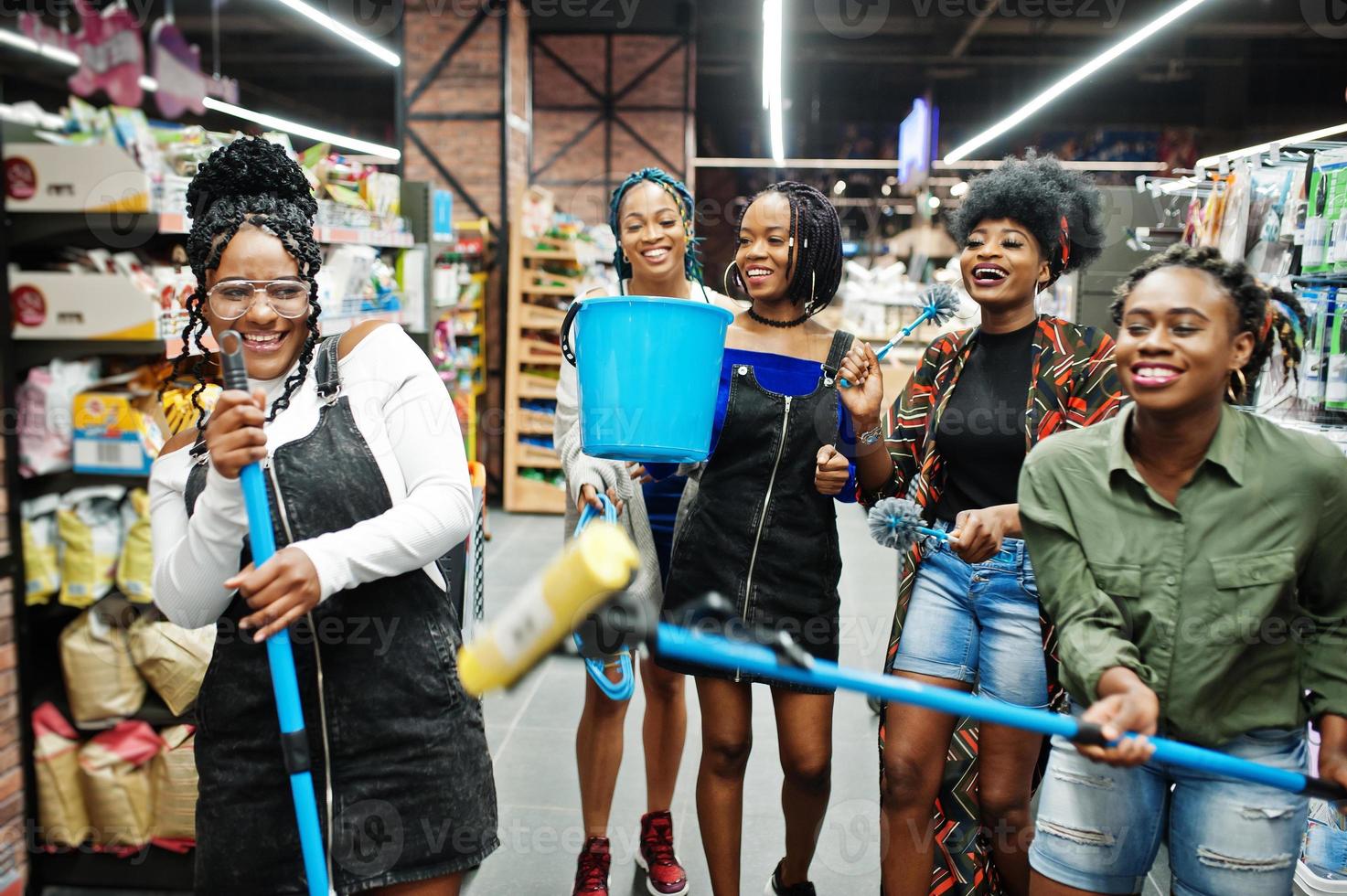 Group of five african womans with dust mop, toilet brush and bucket having fun in household cleaning items department in supermarket. photo