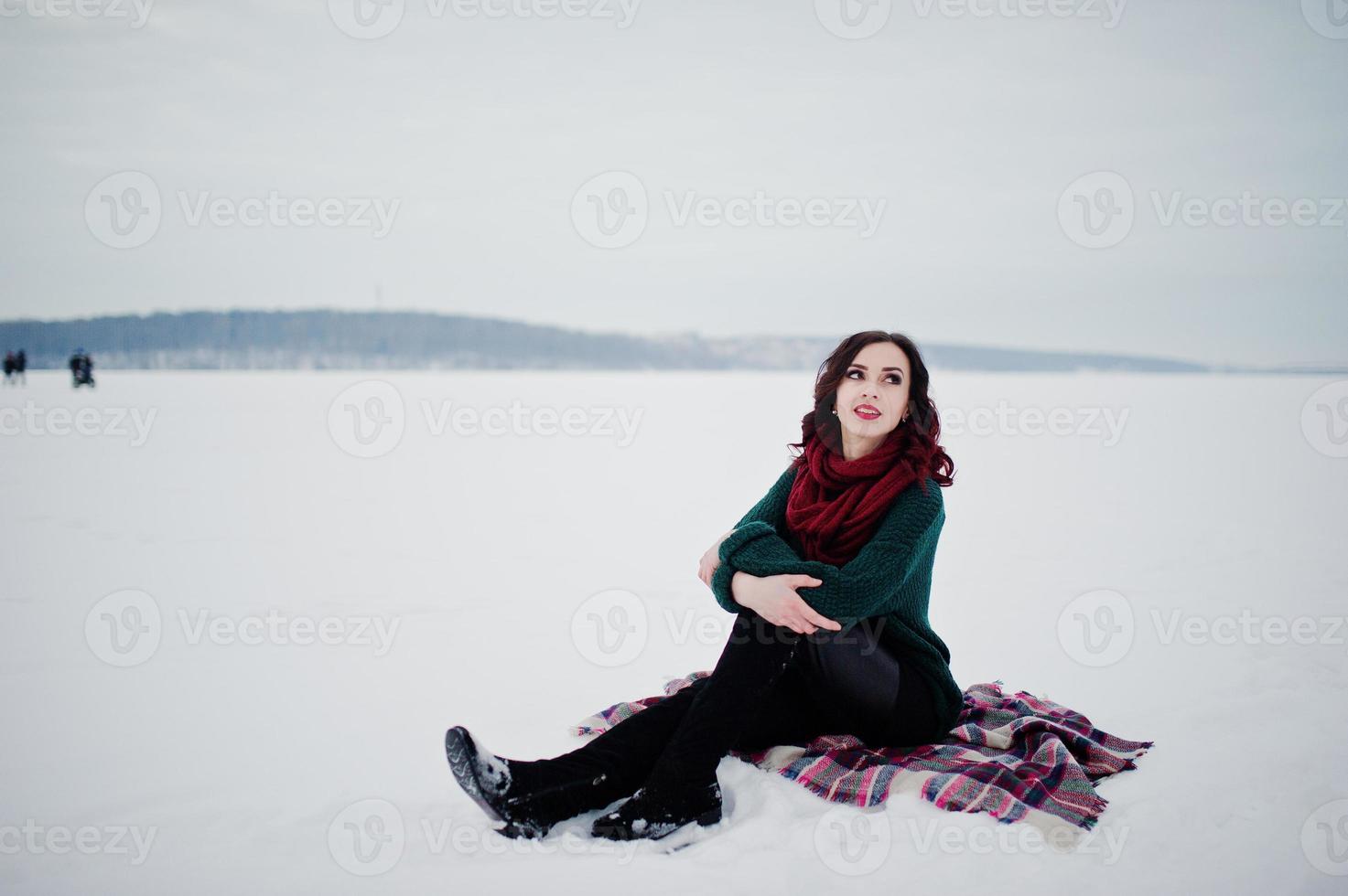 Brunette girl in green sweater and red scarf sitting on plaid outdoor frozen lake on evening winter day. photo