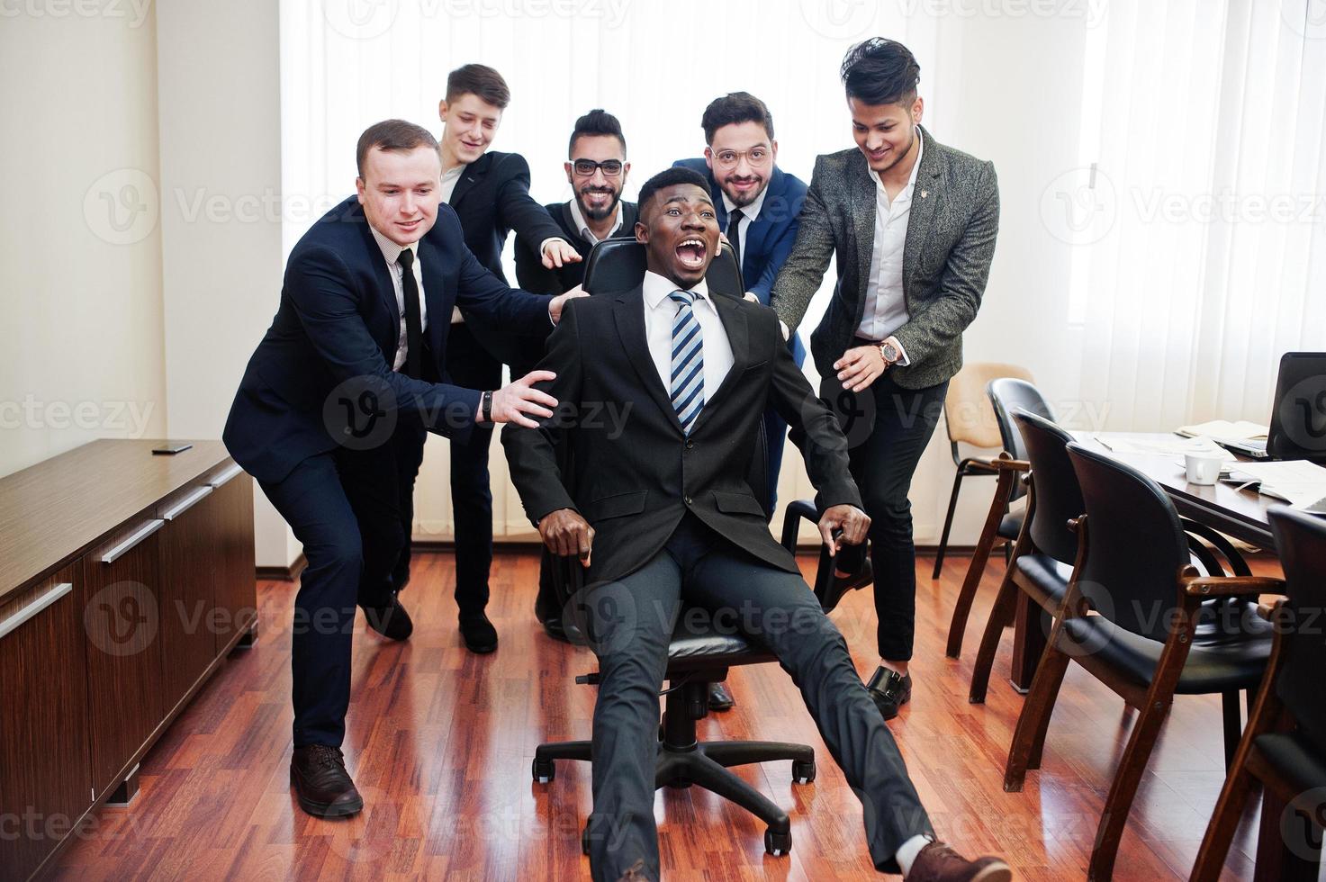 Six multiracial business mans standing at office and roll man on chair. Diverse group of male employees in formal wear having fun. photo