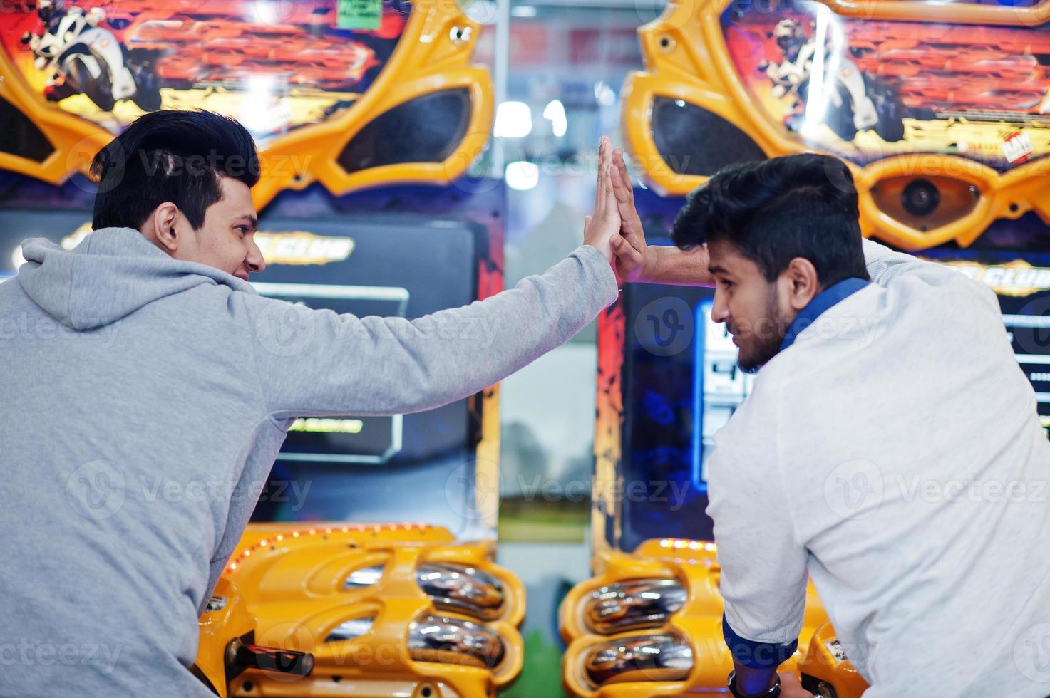 Two asian guys compete on speed rider arcade game motorcycle racing simulator machine. Give high five each other. photo