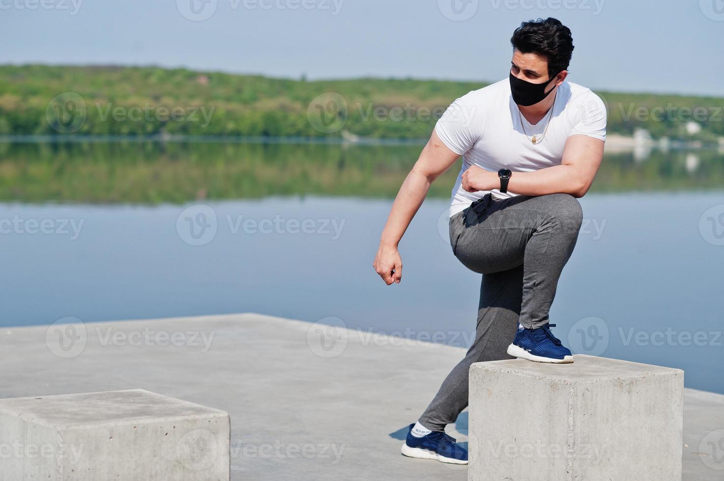 Portrait sports arabian man in black medical face mask doing morning workout exercises against lake during coronavirus quarantine. photo