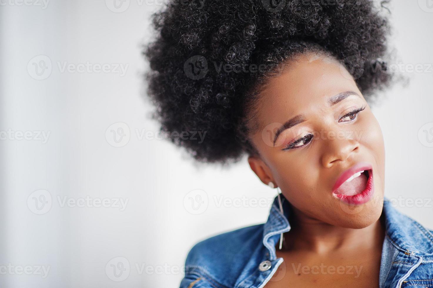 Close up portrait of an attractive african american woman with afro hair wear jeans jacket, posed at white room. Fashionable black model. photo