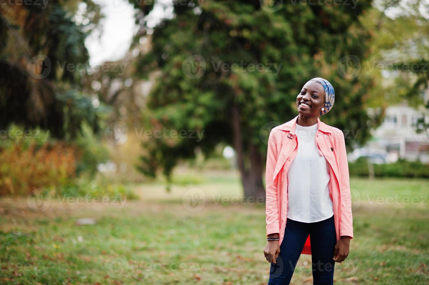 Young modern fashionable, attractive, tall and slim african muslim woman in hijab or turban head scarf and pink coat posed at park. photo
