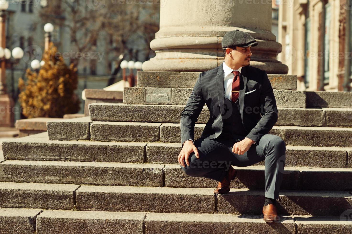 Portrait of retro 1920s english arabian business man wearing dark suit, tie and flat cap near old columns. photo