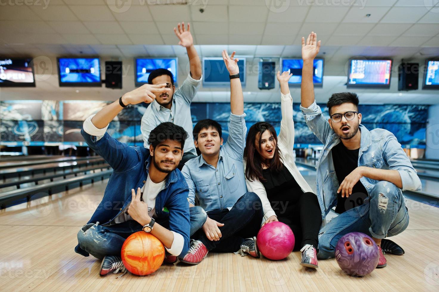 Group of five south asian peoples having rest and fun at bowling club, sitting on bowling alley with balls on hands. photo