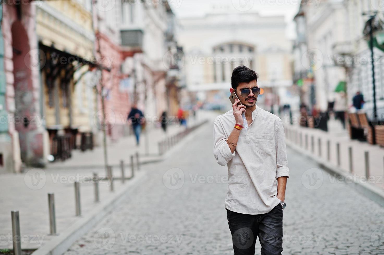 Stylish indian model man in casual clothes and sunglasses posed outdoor at street of India and speaking on mobile phone. photo