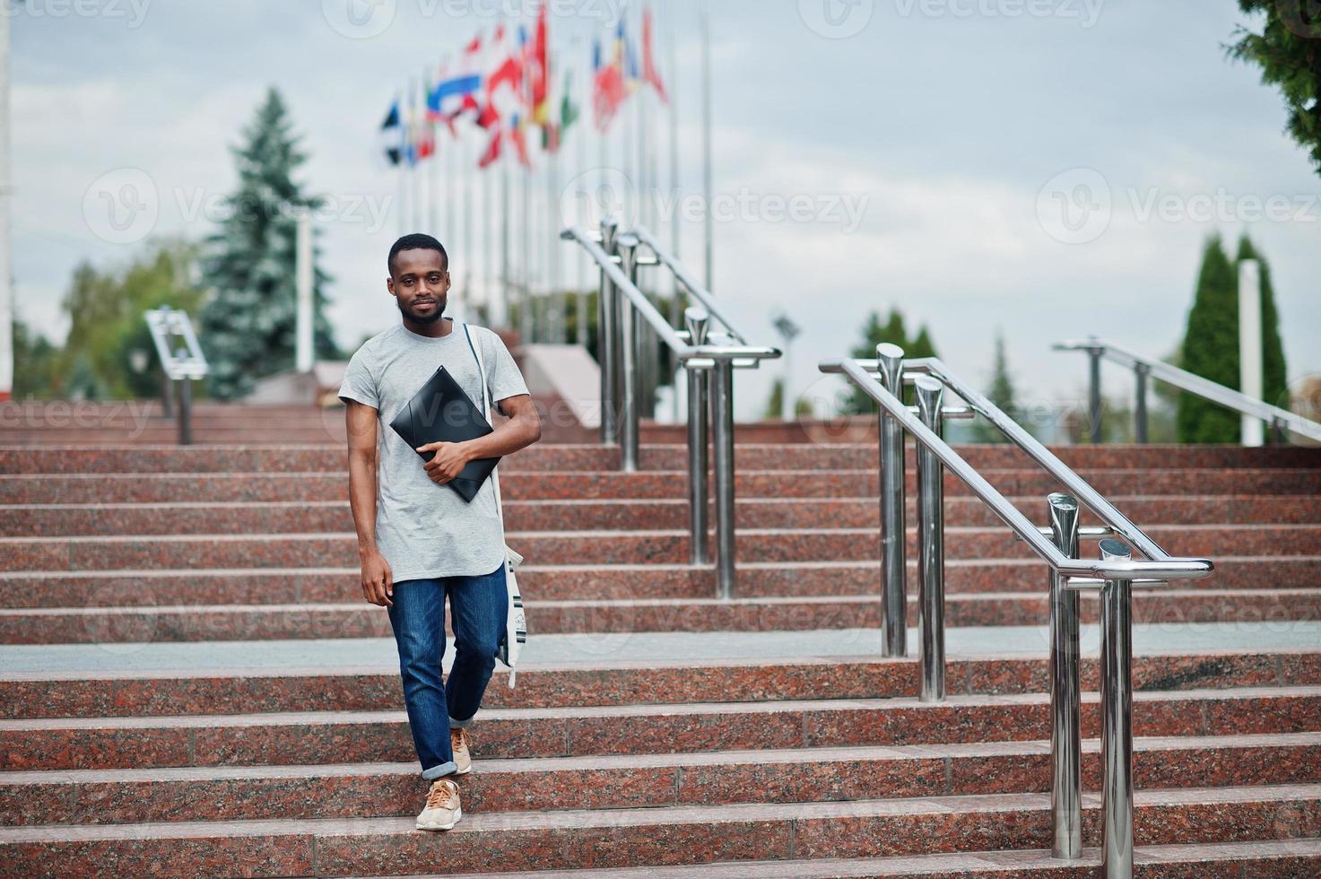 African student male posed with backpack and school items on yard of university, against flags of different countries. photo