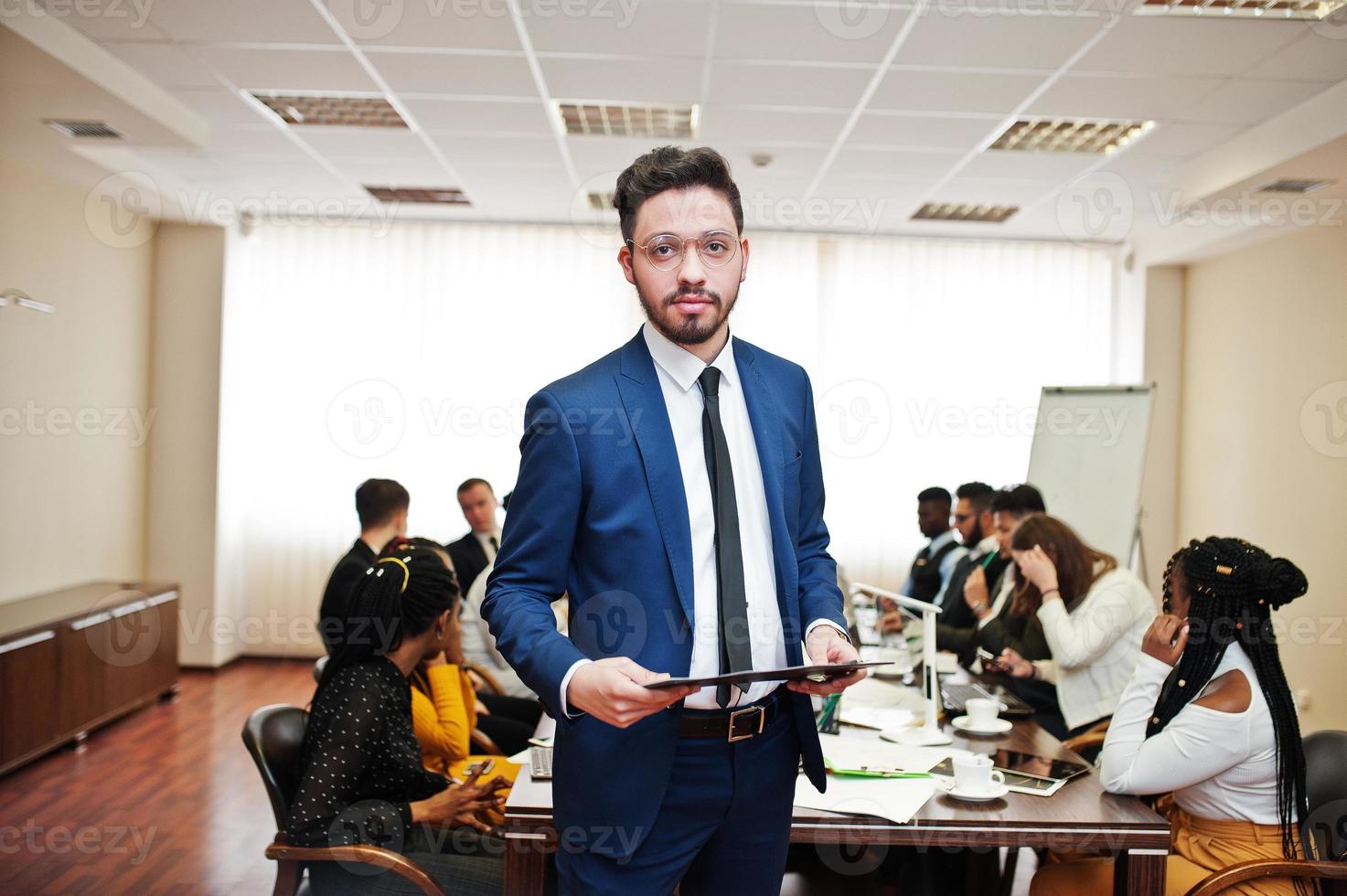 Face of handsome arabian business man, holding clipboard on the background of business peoples multiracial team meeting, sitting in office table. photo