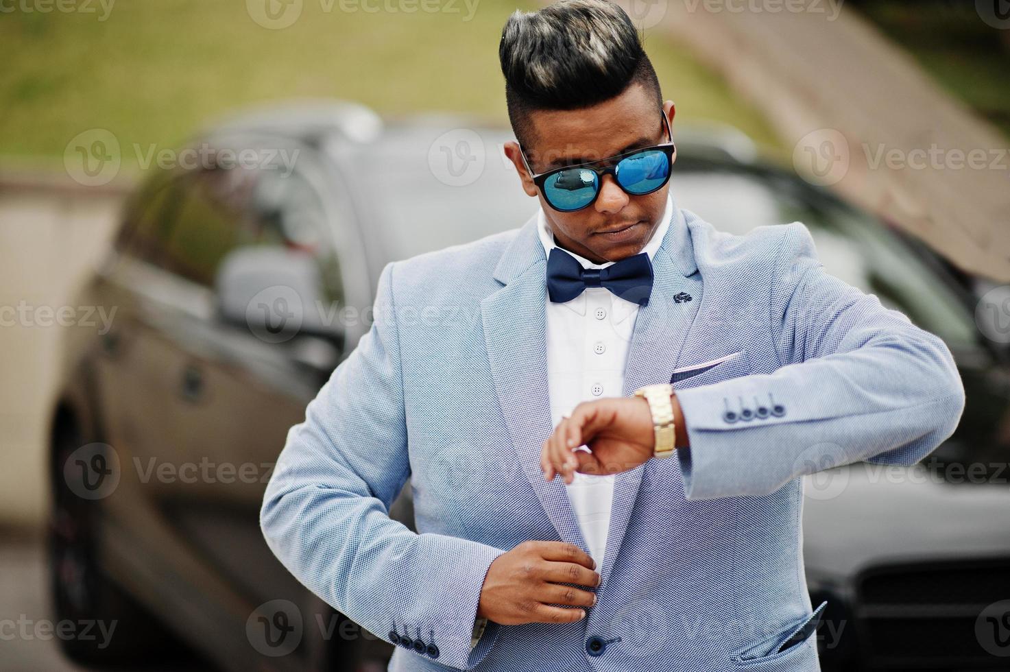 Stylish arabian man in jacket, bow tie and sunglasses against black suv car. Arab rich businessman looking on watches. photo