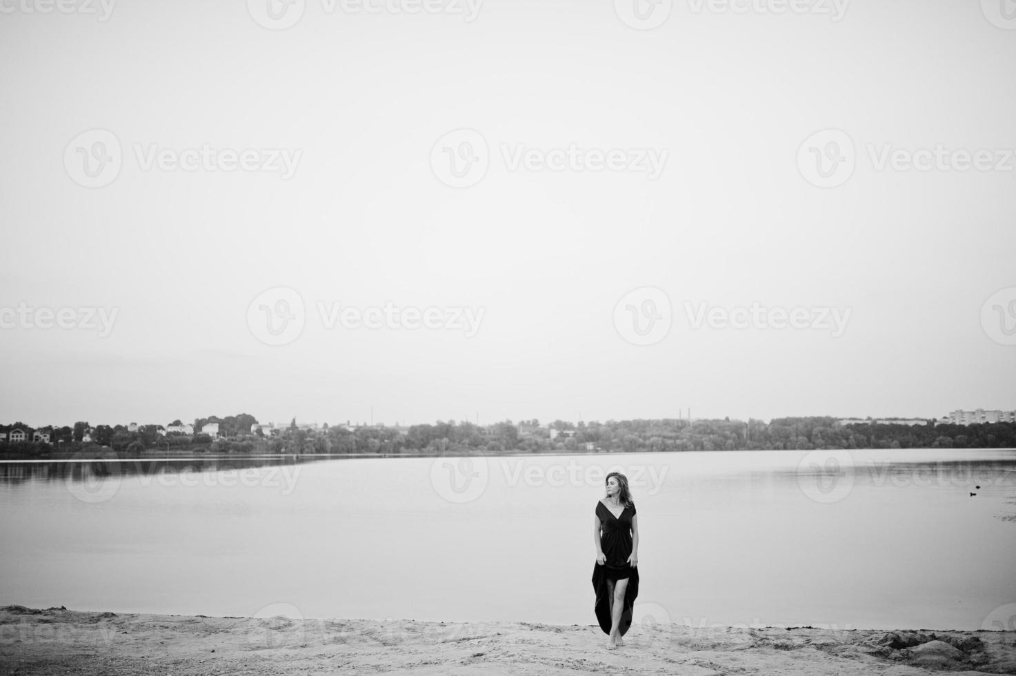 Blonde sensual barefoot woman in red marsala dress posing against lake on sand. photo