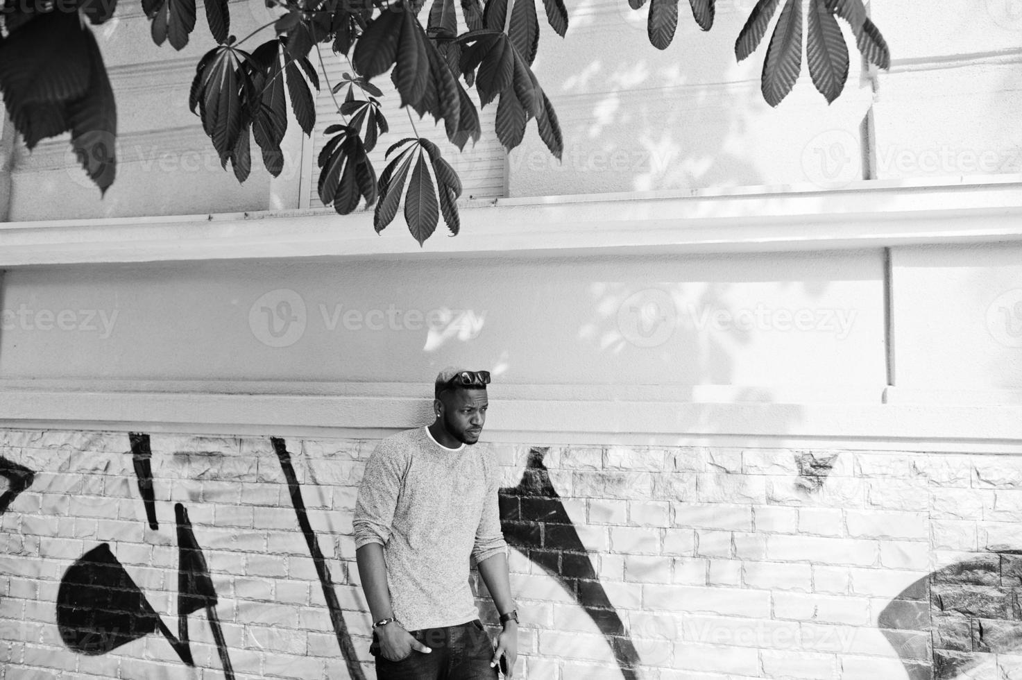 Stylish african american boy on gray sweater and black sunglasses posed at street. Fashionable black guy against graffity wall. photo