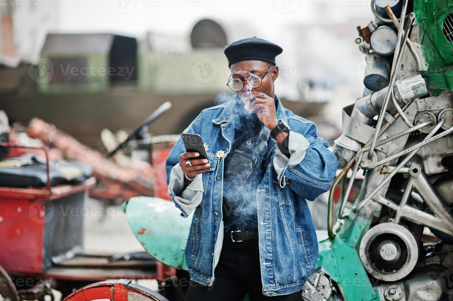 African american man in jeans jacket, beret and eyeglasses, smoking cigar and posed against old retro vehicle and looking at phone. photo