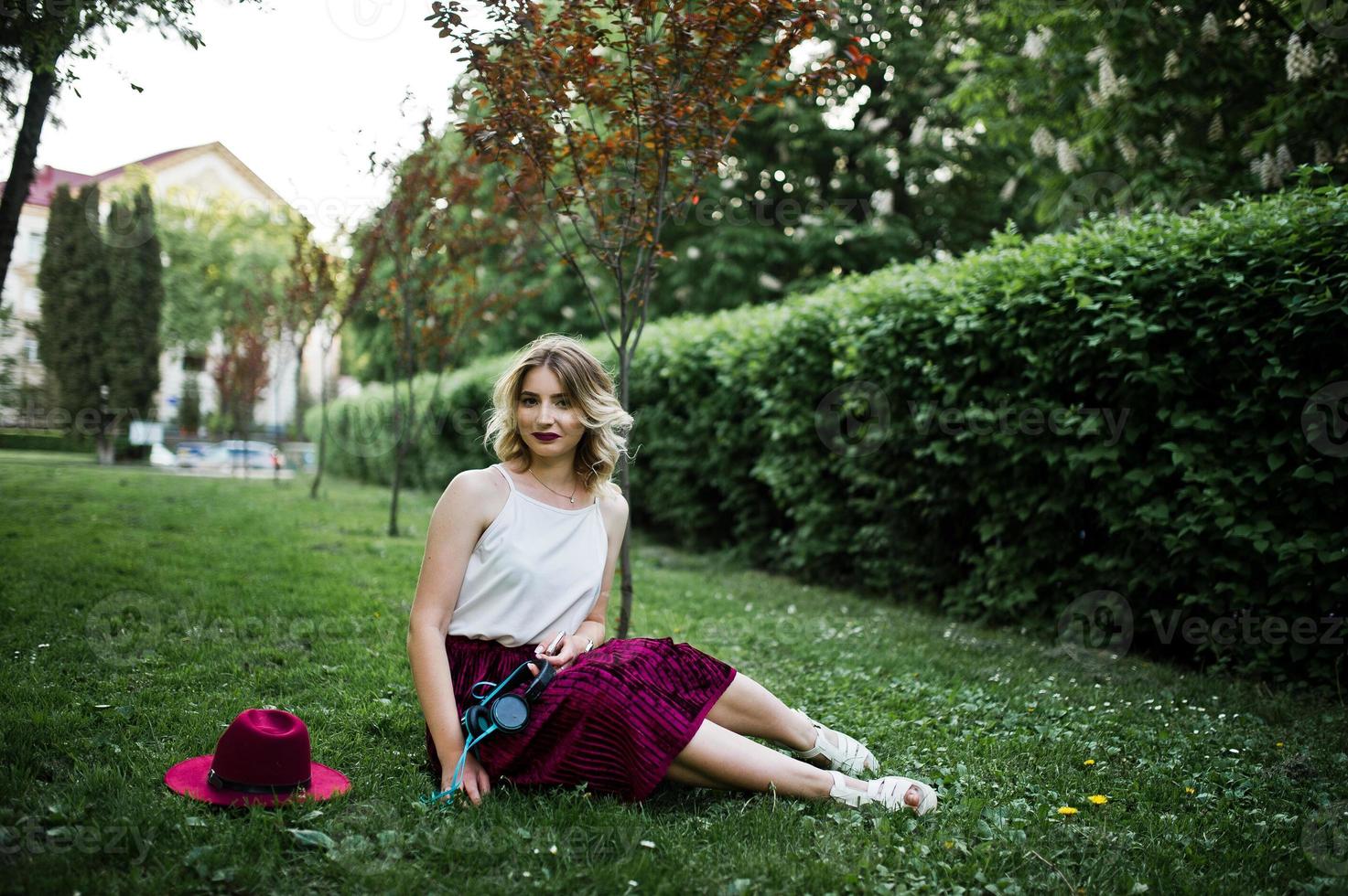 Fashionable and beautiful blonde model girl in stylish red velvet velour skirt, white blouse and hat, sitting on green grass at park with phone and earphones. photo