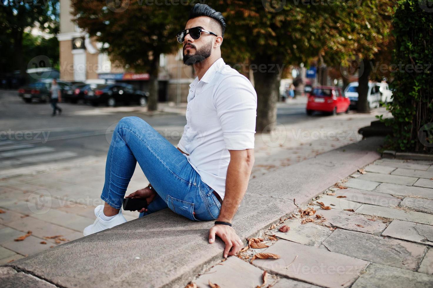 Stylish tall arabian man model in white shirt, jeans and sunglasses sitting at street of city. Beard attractive arab guy at park. photo