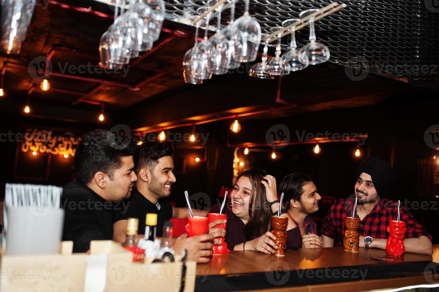 Group of indian friends having fun and rest at night club, drinking cocktails near bar counter. photo