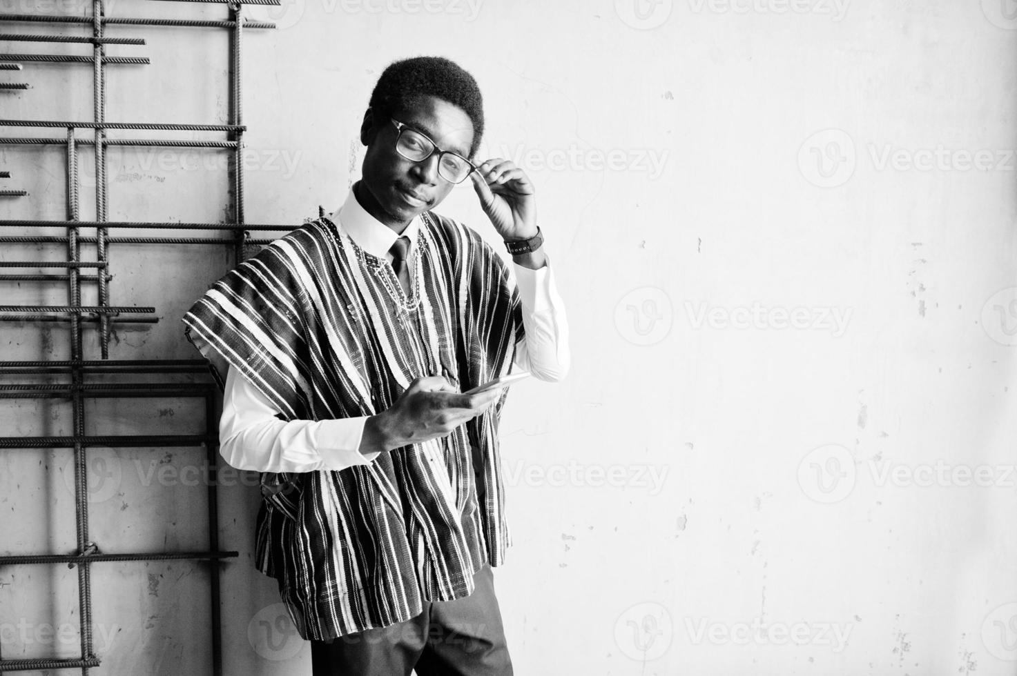 African businessman in traditional clothes and glasses with mobile phone at hand posed at modern building indoor. photo