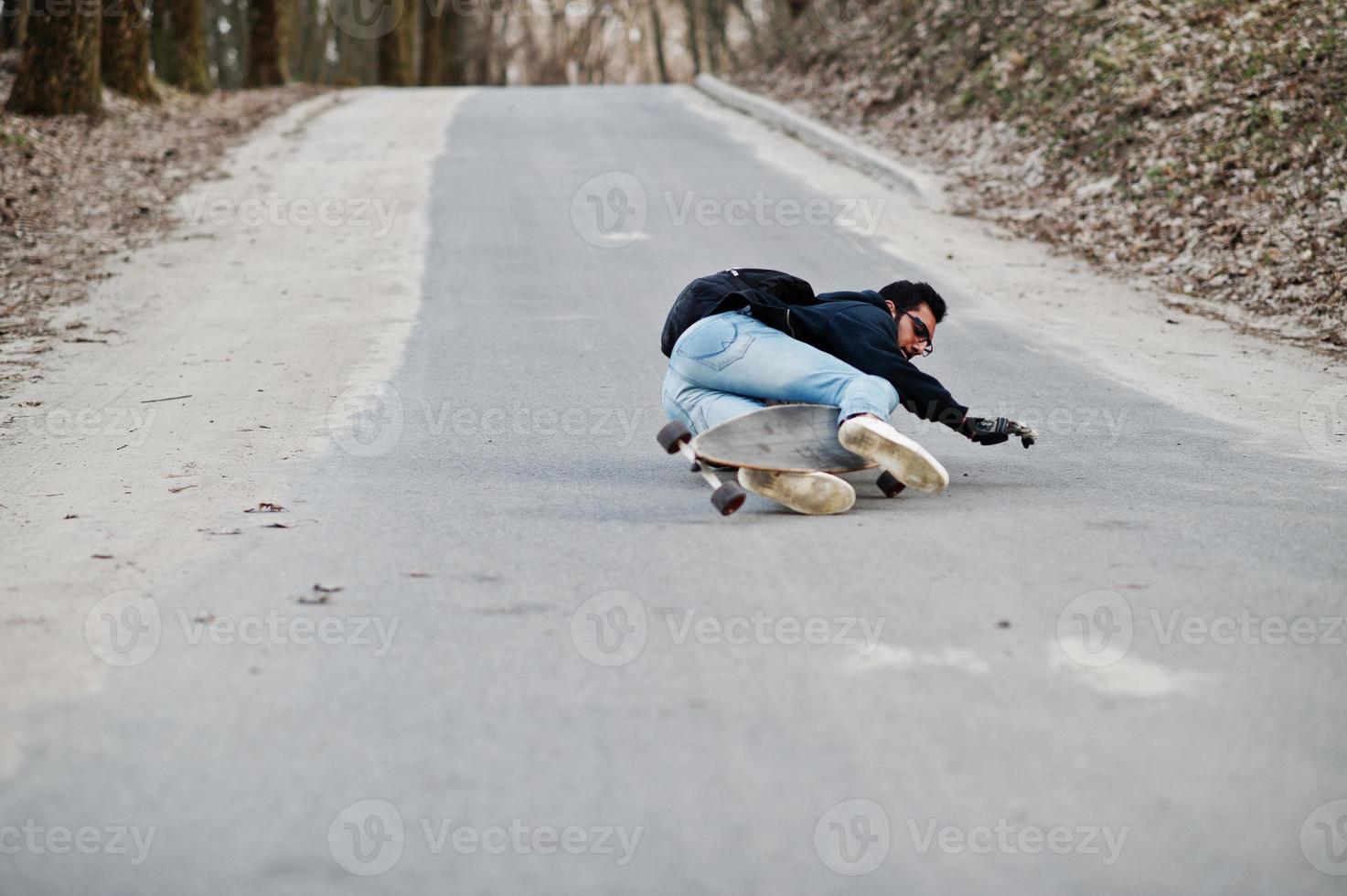 fallar al caer de una patineta. hombre árabe de estilo callejero en anteojos con longboard longboarding por el camino. foto