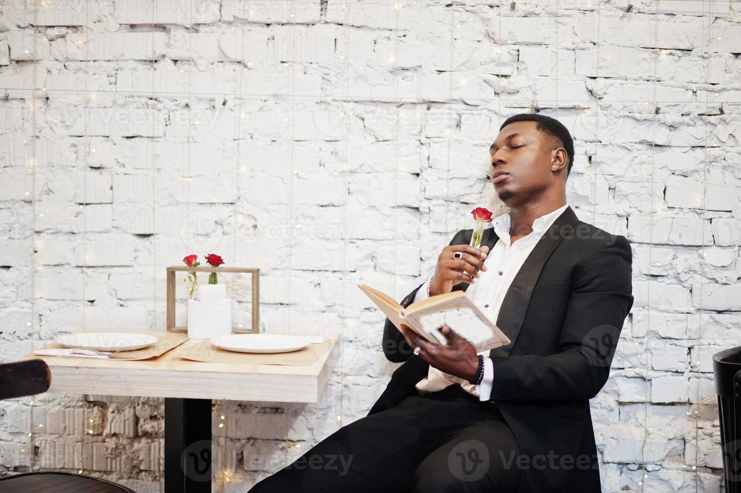 Strong powerful african american man in black suit sitting at cafe, read book and sniff flower in test tube. photo