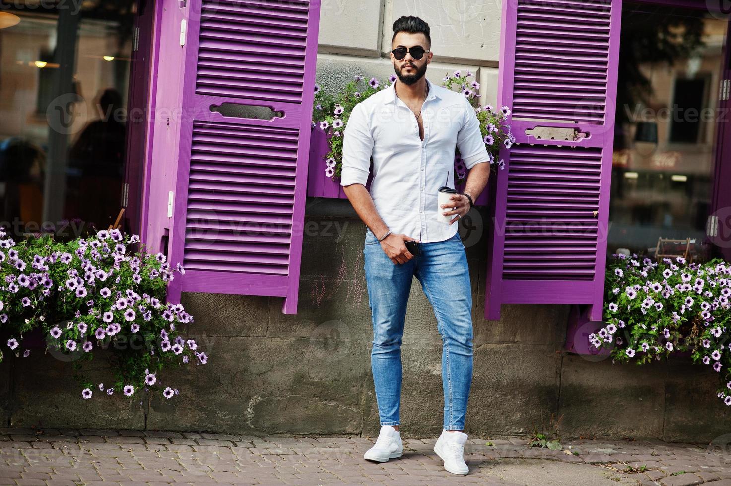 Stylish tall arabian man model in white shirt, jeans and sunglasses posed at street of city. Beard attractive arab guy with cup of coffee against purple windows. photo