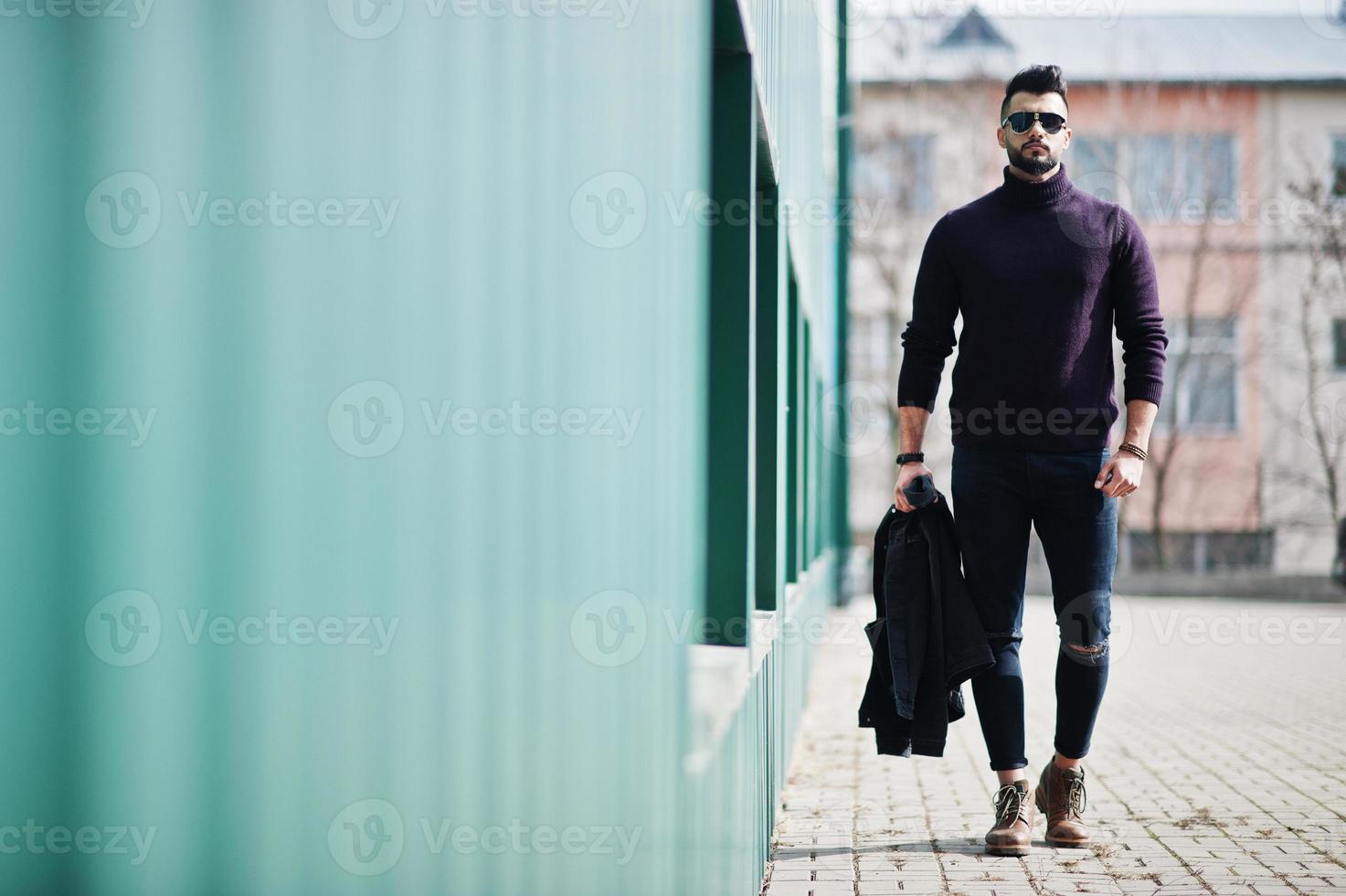 Fashion rich beard Arab man wear on turtle neck and sunglasses walking against green wall with jacket on hand. Stylish, succesful and fashionable arabian model guy. photo