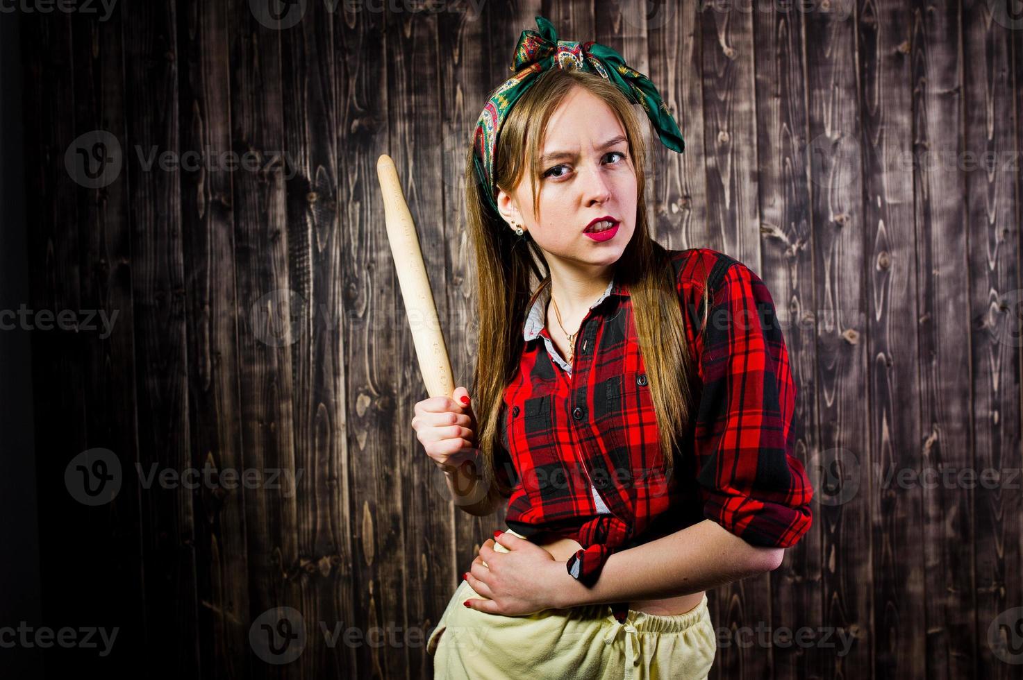 Young funny housewife in checkered shirt and yellow shorts pin up style with kitchen rolling pin on wooden background. photo