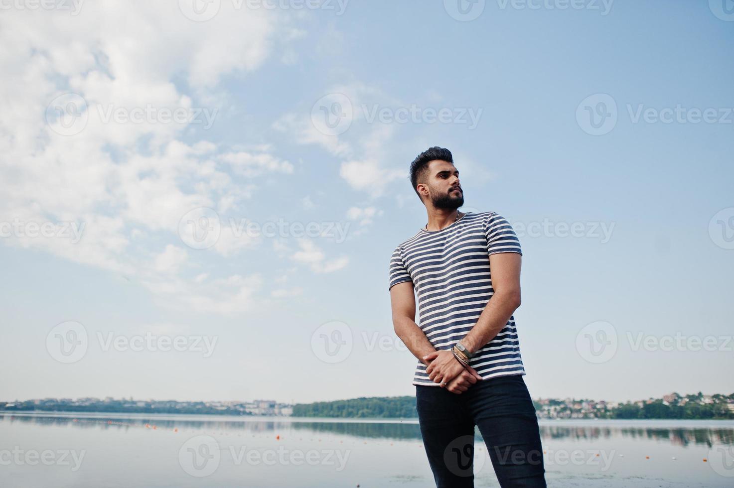 Handsome tall arabian beard man model at stripped shirt posed outdoor against lake and sky. Fashionable arab guy. photo